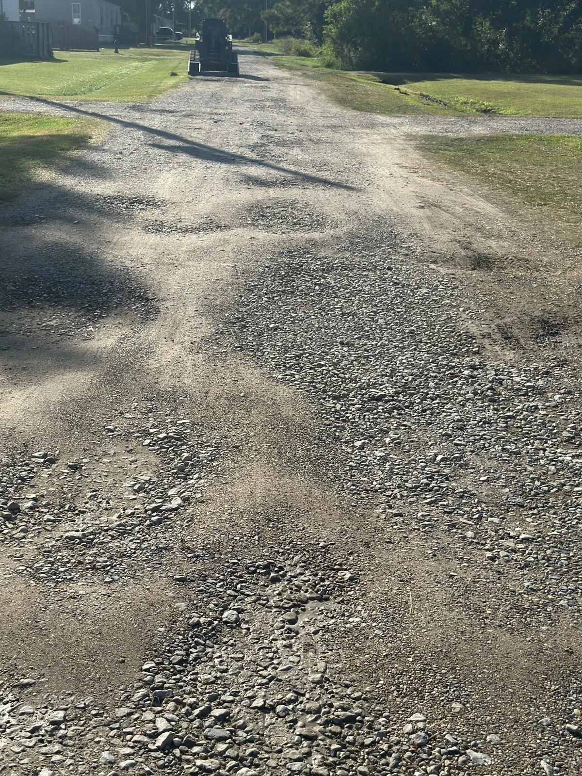 Potholed gravel driveway in south Louisiana before regrading, with a compact track loader working at the far end.