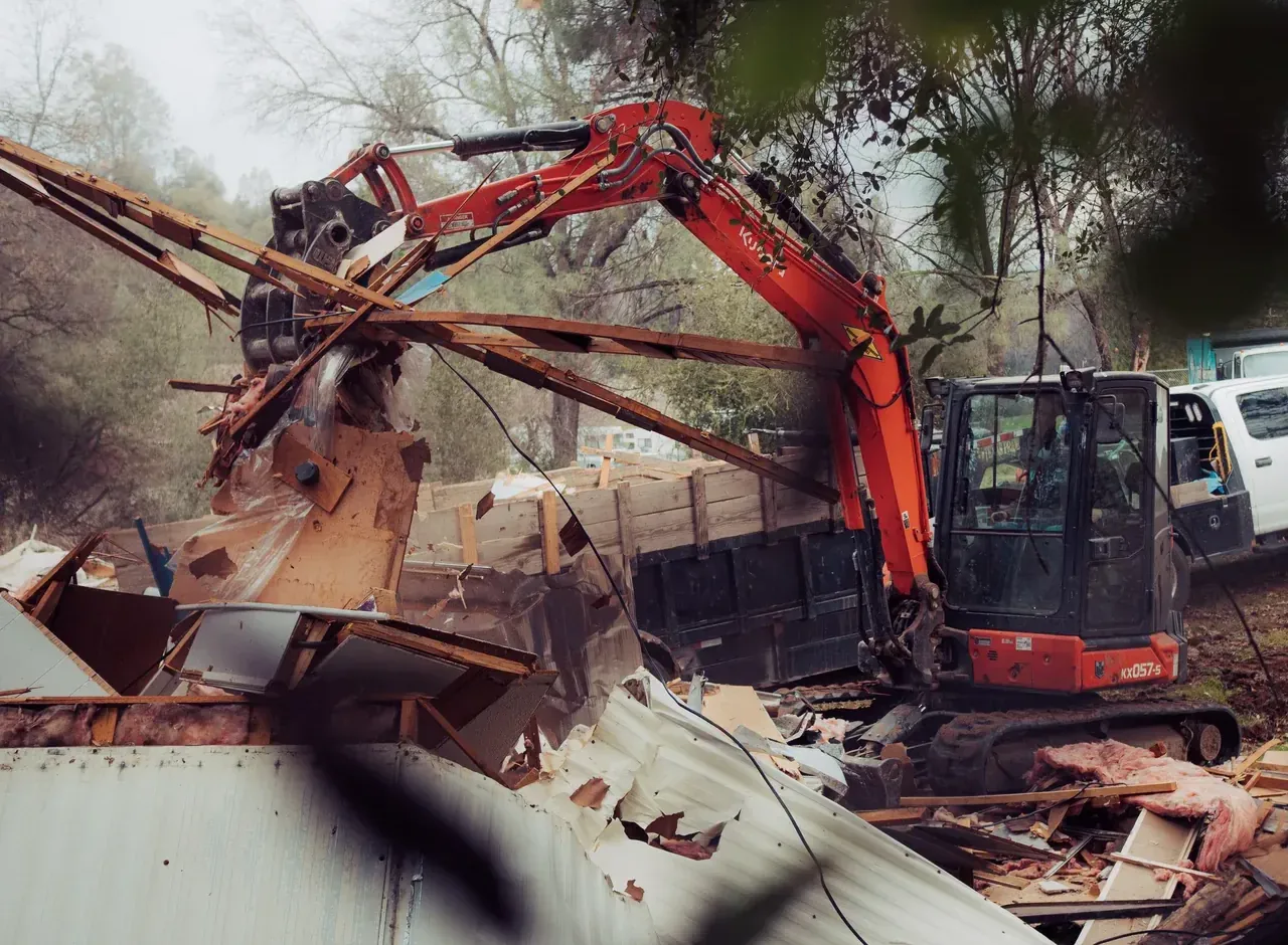 Mini excavator tearing apart an old mobile home, with a dump trailer behind it
