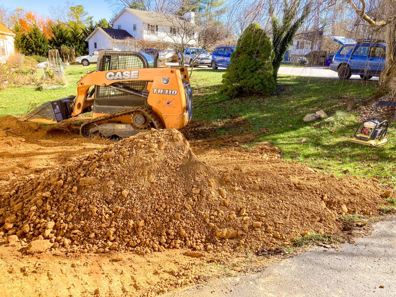 Compact track loader spreading a pile of fill across a yard, with a plate compactor on the lawn